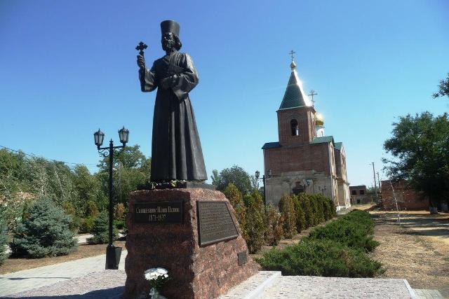 Общий вид памятника о. Илие Попову на фоне храма мучеников Флора и Лавра
